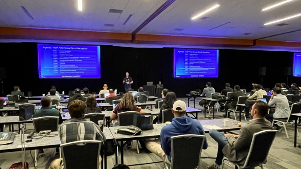 Developers at laptops in a conference hall during the Intel AI for Social Good hackathon, with the agenda on screens up front